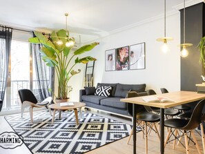 Un salon moderne avec un canapé gris, un tapis à motifs noir et blanc, une table basse en bois et un coin repas avec une table en bois et des chaises noires. On y trouve une grande plante en pot et un luminaire suspendu au-dessus du canapé.