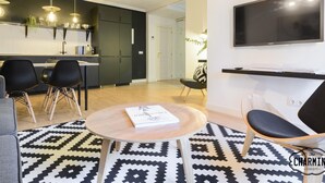 Ein modernes Wohnzimmer mit einem runden Holztisch, einem schwarz-weißen Teppich, einem grauen Sofa und einem schwarzen Sessel. Es gibt eine Küchenzeile mit dunklen Schränken und einem integrierten Fernseher über der Arbeitsfläche.