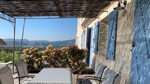 Um pátio externo com mesa e cadeiras, uma área coberta com telhado de palha, e vista para colinas ao fundo.