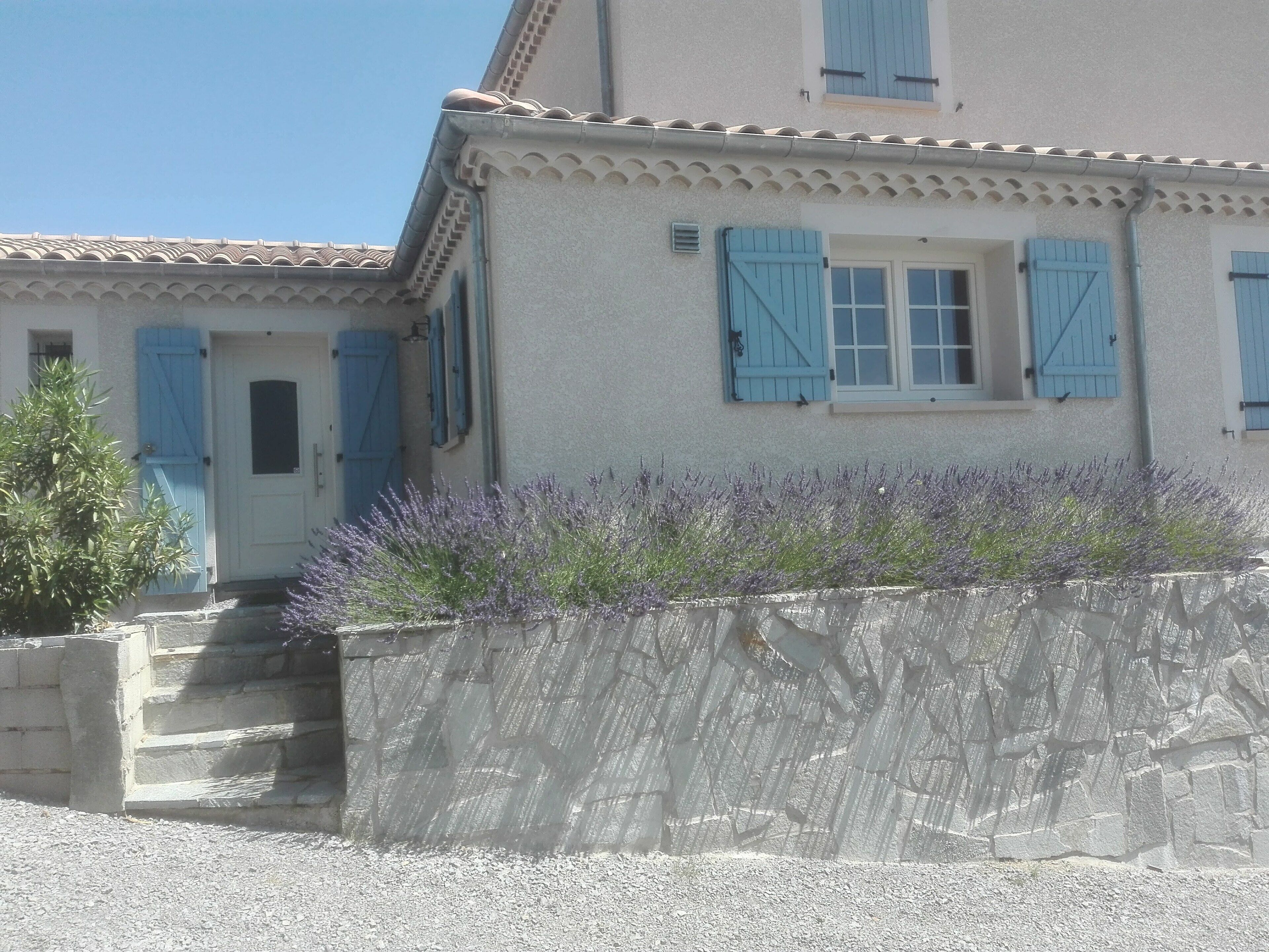 Una casa a due piani con persiane blu, una porta bianca e una piccola finestra. Si apre su un vialetto sterrato e un muro in pietra con piante di lavanda.