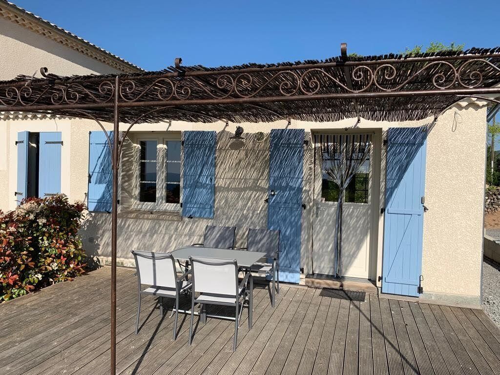 Een terras met tafel en stoelen, een pergola, en een gebouw met blauwe luiken.
