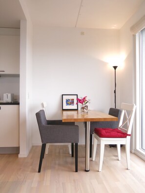 Ett modernt matområde med ett träbord, två grå stolar och en vit stol med en röd kudde. Ett inramat fotografi och en vas med blommor står på bordet.