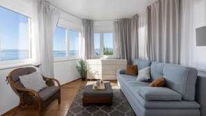 Una sala de estar moderna con un sofá, una silla, una mesa de centro de madera y vista al mar a través de grandes ventanales.
