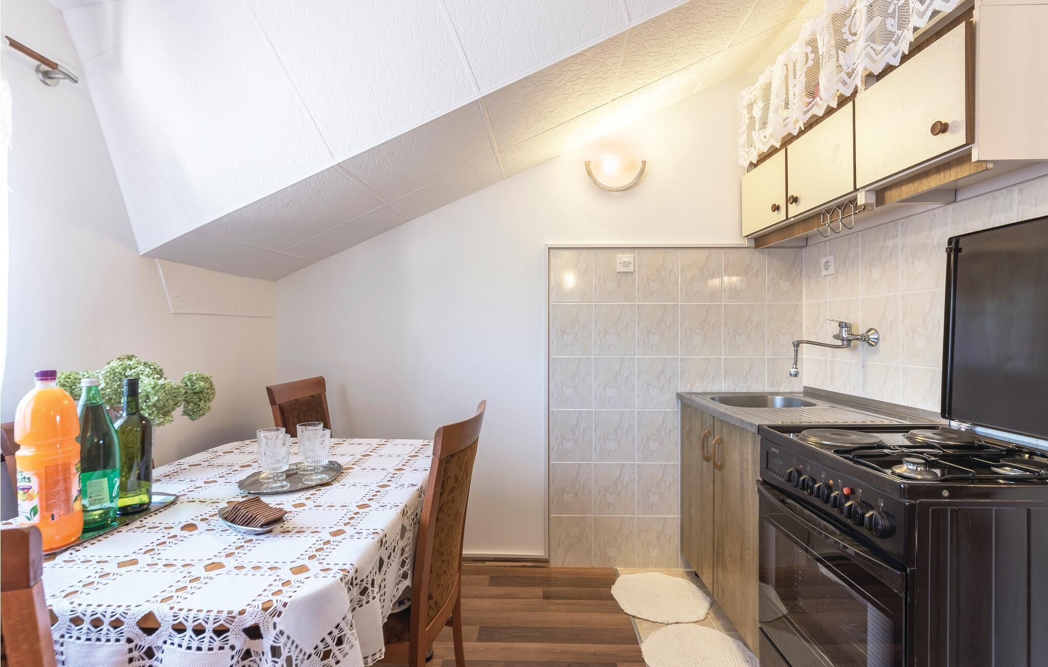 Een kleine keuken met een fornuis, spoelbak en een betegelde spatwand. Er is een eettafel met een kanttafelkleed en stoelen.