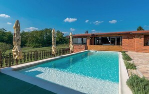 Una piscina rectangular con un deck de madera y una casa de ladrillo al fondo.