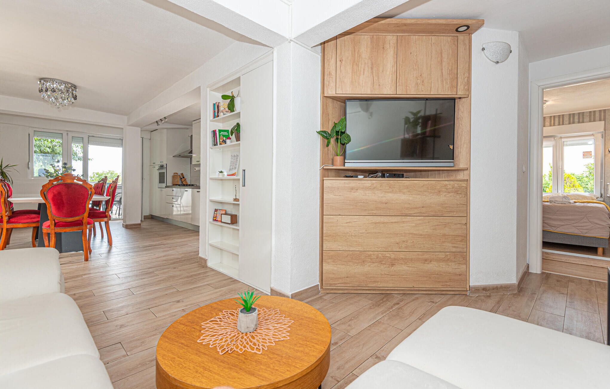 Uma sala de estar moderna com um móvel de televisão de madeira, uma área de jantar com cadeiras vermelhas e uma cozinha ao fundo.