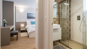 Ett modernt hotellrum med en säng, ett nattduksbord och en dusch som är avskärmad med glasväggar.