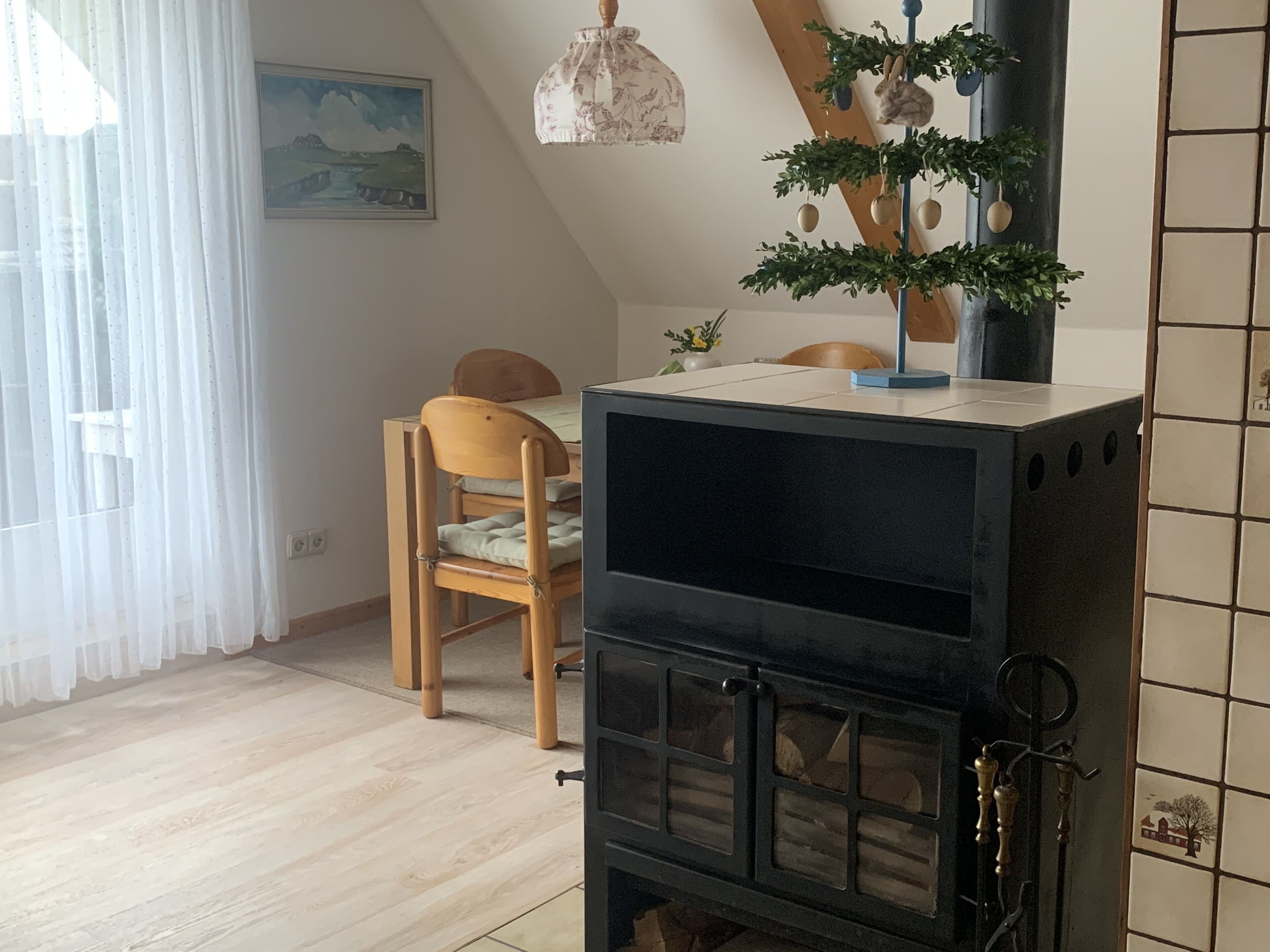 Ein gemütliches Zimmer mit einem Holzstuhl, einem Tisch, einem schwarzen Herd und einem Fenster mit durchsichtigen Vorhängen.