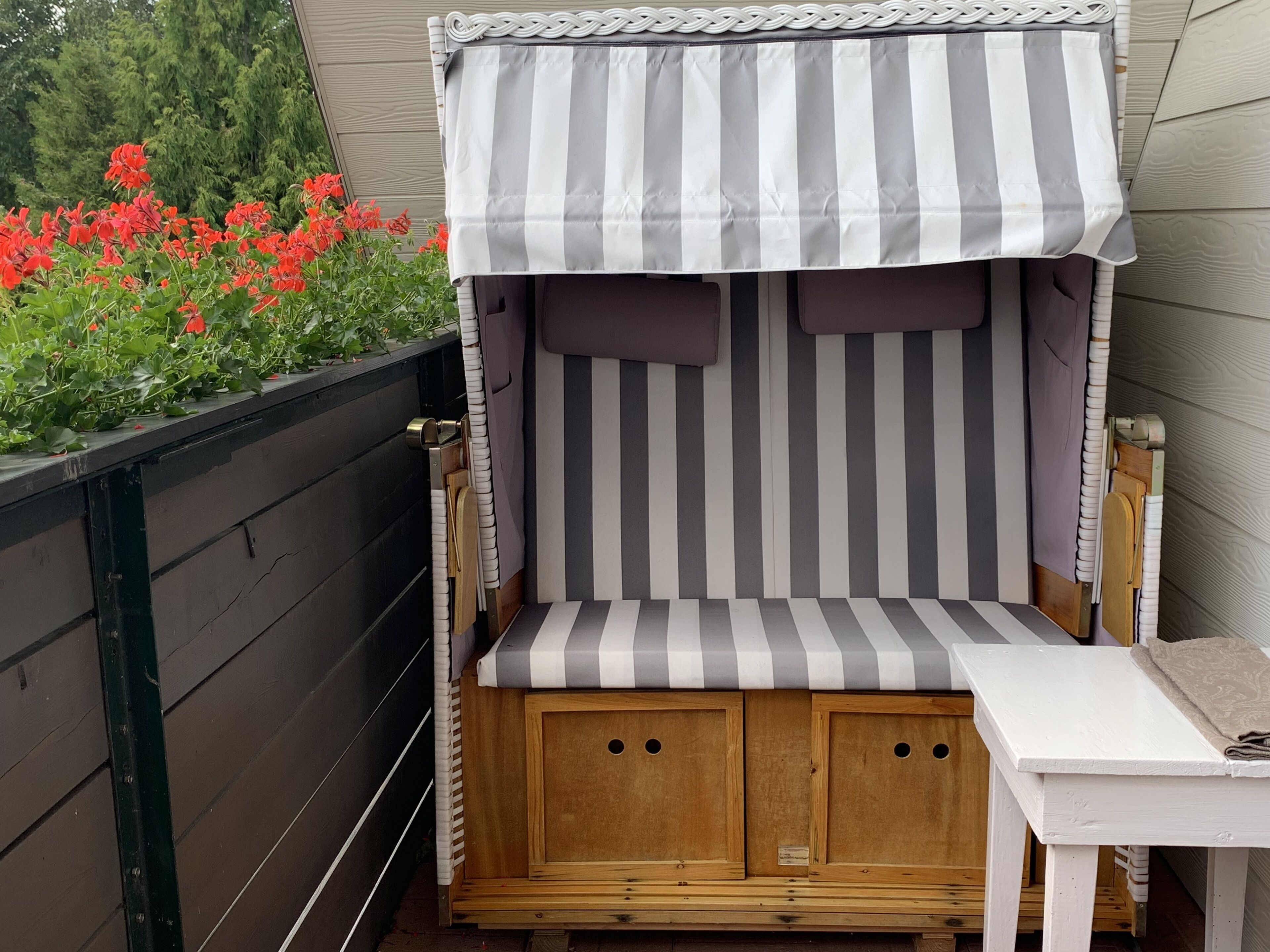 Een kleine houten bank met een gestreept zonnescherm, een tafel en een houten omheining met rode bloemen.