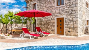 A stone building with a pool, red umbrellas, and lounge chairs.