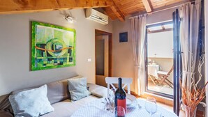 Een gezellige kamer met een houten plafond, een schilderij aan de muur, een eettafel gedekt met een fles, glazen en borden, en uitzicht op een terras.