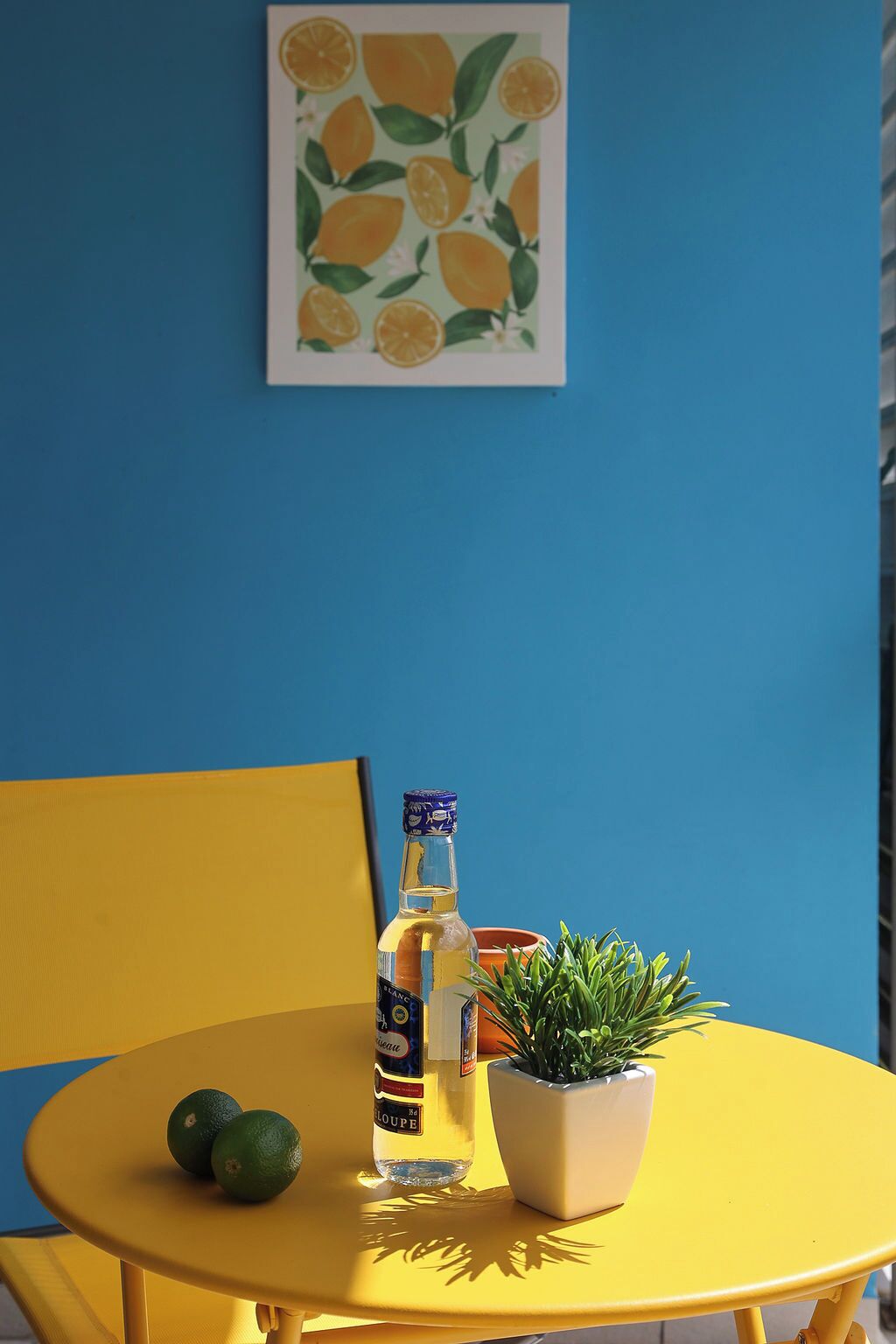 A yellow table with a bottle of white wine, two limes, and a potted plant against a blue wall with a lemon-themed artwork.