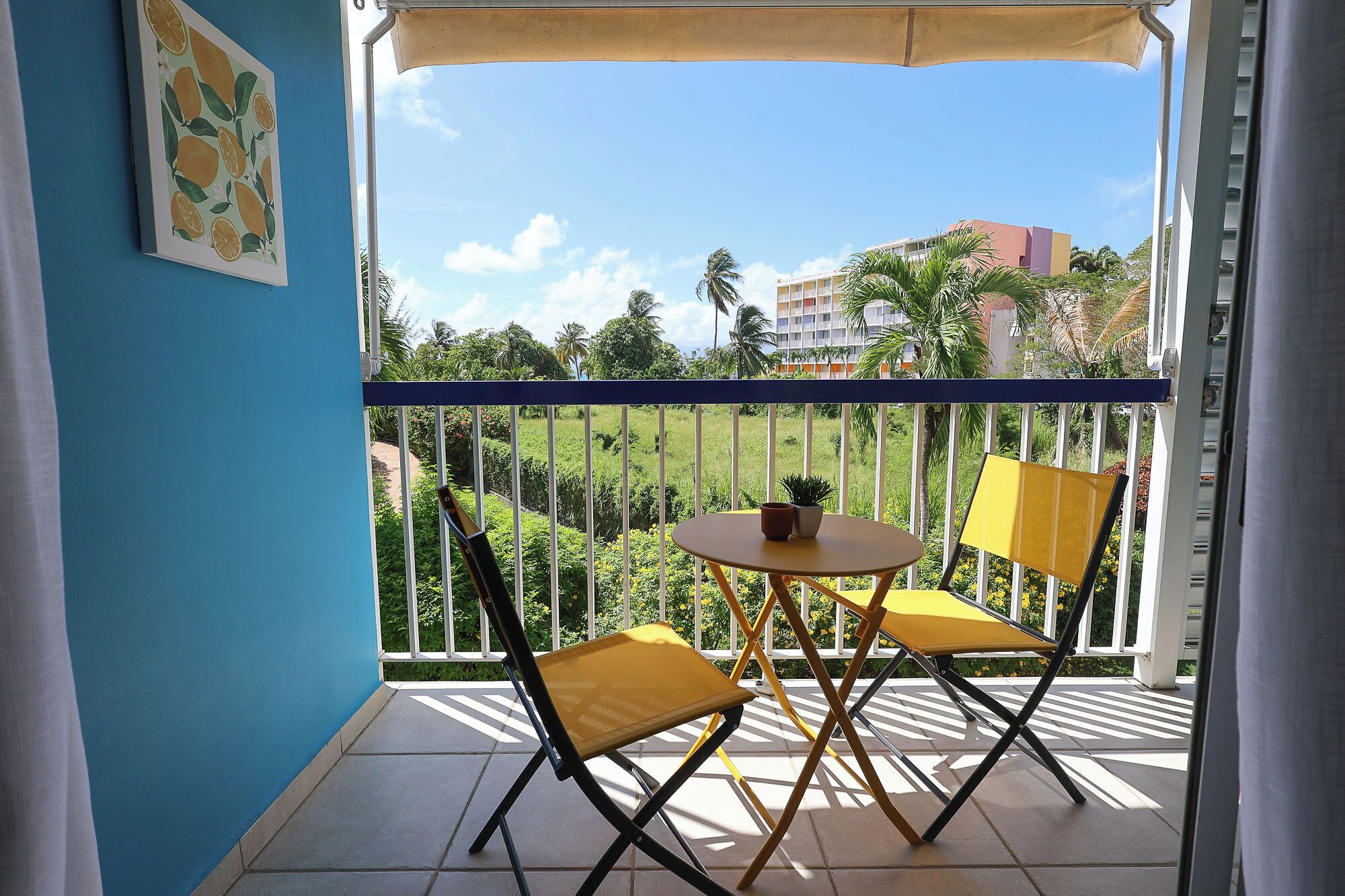 Un balcone con due sedie gialle, un tavolino con una pianta e un quadro appeso al muro.