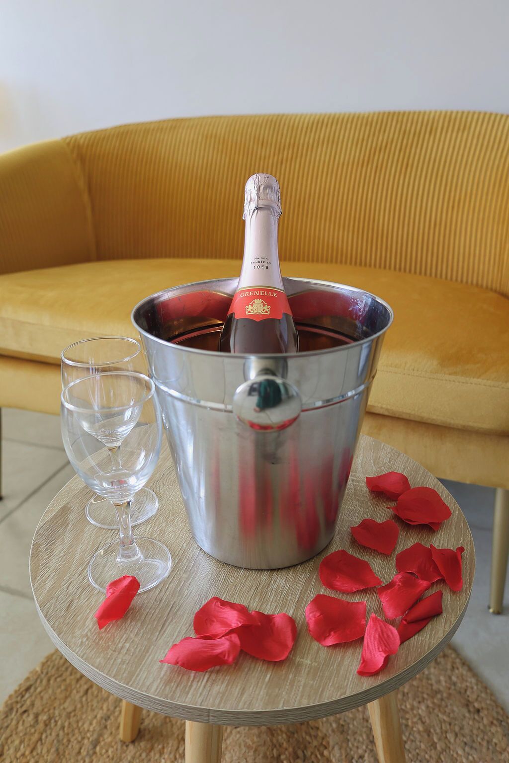 A bottle of Grenelle champagne in a metal bucket, a glass, and rose petals on a small round table.