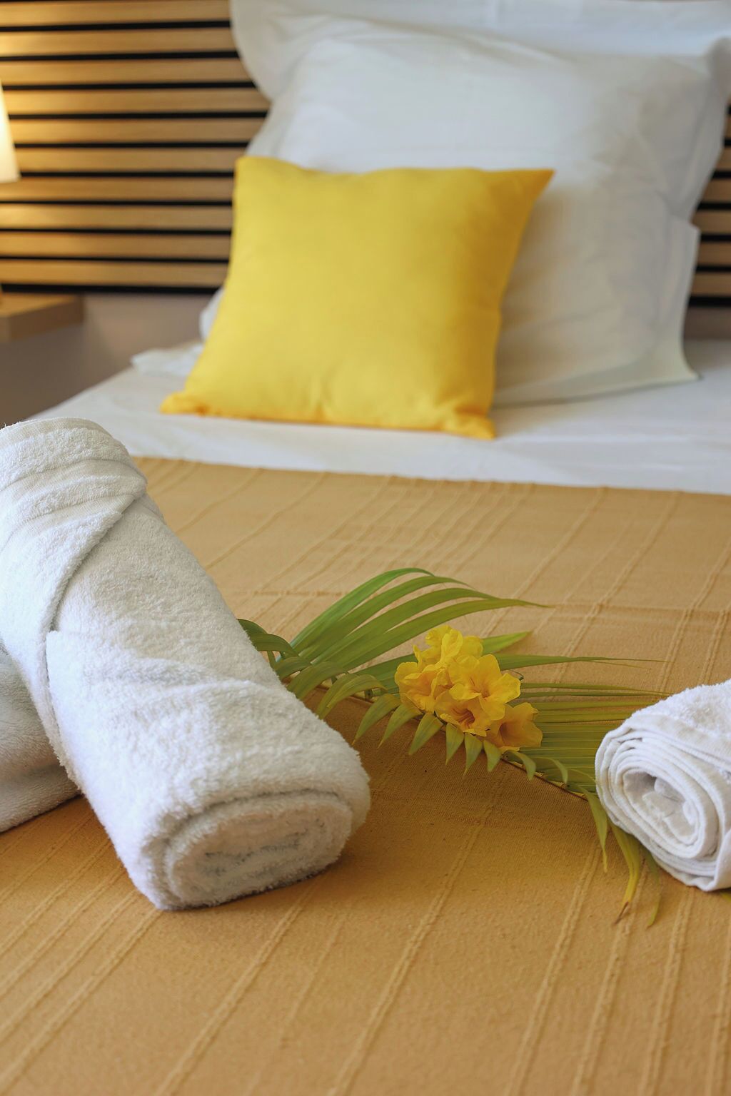 A neatly made bed with white and yellow pillows, a rolled white towel, and a bouquet of yellow flowers on a wooden floor.
