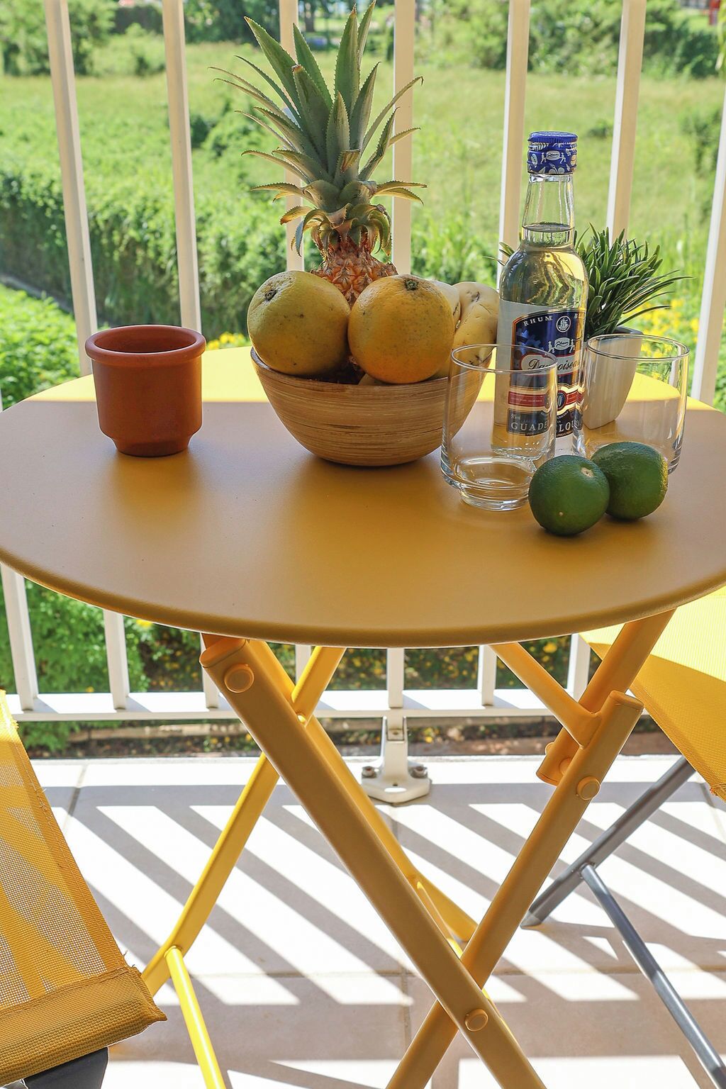 A round yellow table with a bowl of fruits, a glass, and a bottle on a balcony with a view of greenery.