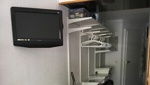 Habitación compacta con escritorio blanco, monitor negro, armario blanco y estantería con ropa colgada.