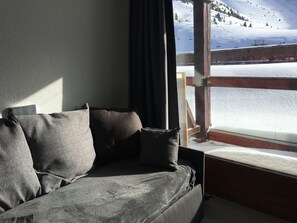 Ein Zimmer mit grauem Sofa und Fenster mit Blick auf eine verschneite Landschaft und eine Seilbahn.