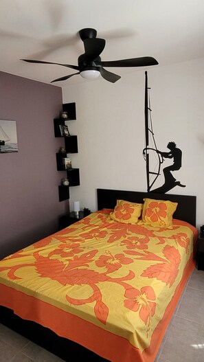 Un dormitorio con cama, ventilador de techo, estante empotrado con libros y un adorno en la pared.