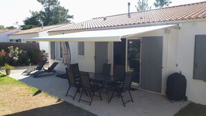 A covered patio with a table and chairs, a garden with flowers, and a house with a tiled roof.