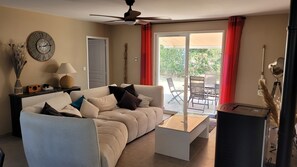 Ein Wohnzimmer mit einer Couch, einem Couchtisch und einem Deckenventilator. Ein Fenster mit roten Vorhängen bietet einen Blick ins Freie.