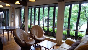 Una habitación con suelo de madera, una mesa redonda, dos sillas, un sofá y ventanas amplias con vista a árboles.