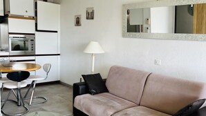 A modern living room with a beige sofa, a glass coffee table, a dining area with a chair, a lamp, and a kitchenette with a refrigerator and microwave. - Apartment in lake city of Grimaud harbor with beautiful view (Port Grimaud)