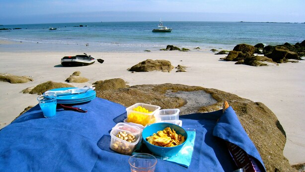 Een strandpicknick met een tafel, borden, bestek en bakjes eten.