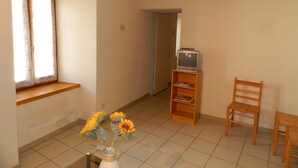 Una habitación con una silla de madera, una mesita pequeña, un televisor sobre un mueble bajo y un jarrón con girasoles.