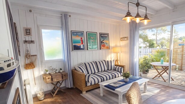 A cozy living room with a striped sofa, a wooden coffee table, and a wicker chair. There are framed pictures on the wall, a lamp, and a view of the outdoors through large windows. - Romantic cottage with private heated pool - 2 pers - Bassin d'Arcachon (Lanton)