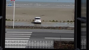 Blick von einem Fenster auf einen Strand, inklusive eines weißen Autos auf der Straße, einer Flagge und einem wolkenlosen Himmel.