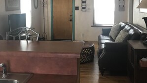 A spacious living room with a wooden door, a ceiling fan, a black leather sofa, and a dining area with a metal chair and table. - LAVENDER  LIZARD B Upper Unit CASITAS (Farmington)
