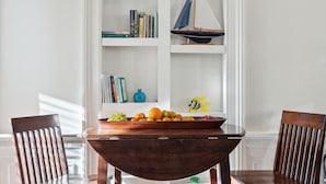 Un coin repas avec une table et des chaises en bois, un panier de fruits et une étagère blanche où sont disposés des objets décoratifs et des livres.