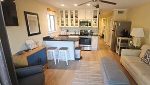 A modern kitchen with white cabinets, a central island with stools, a dining area with a sofa and cushions, and a ceiling fan. - Adorably furnished and updated Ocean Villa condo, with wonderful 200+ Reviews! (Hilton Head Island)