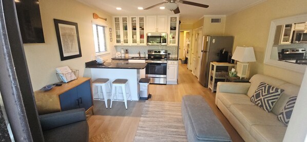 A modern kitchen with white cabinets, a central island with stools, a dining area with a sofa and cushions, and a ceiling fan.