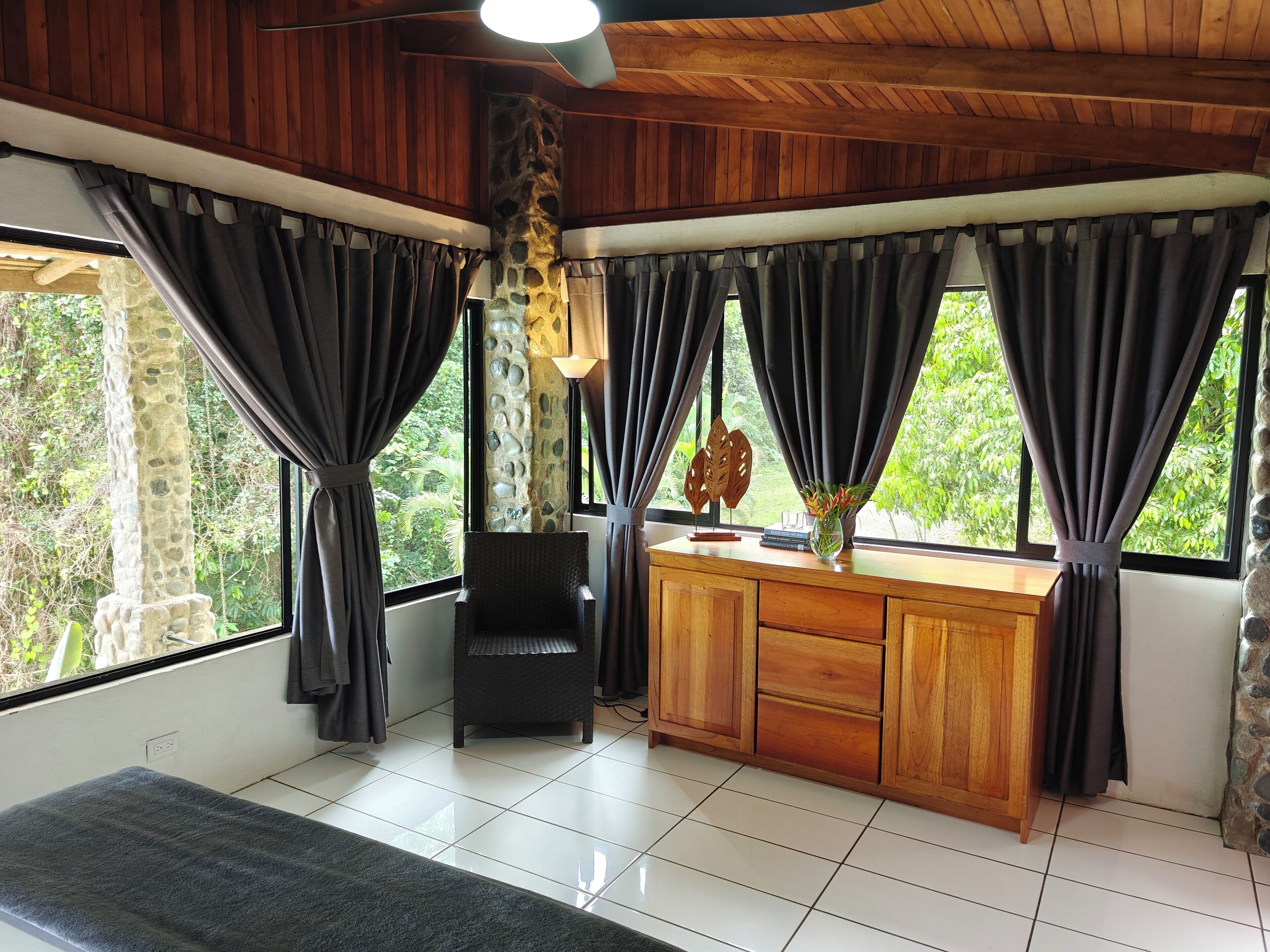 A room with a wooden ceiling, stone pillar, a desk with drawers, a chair, and a window with curtains.