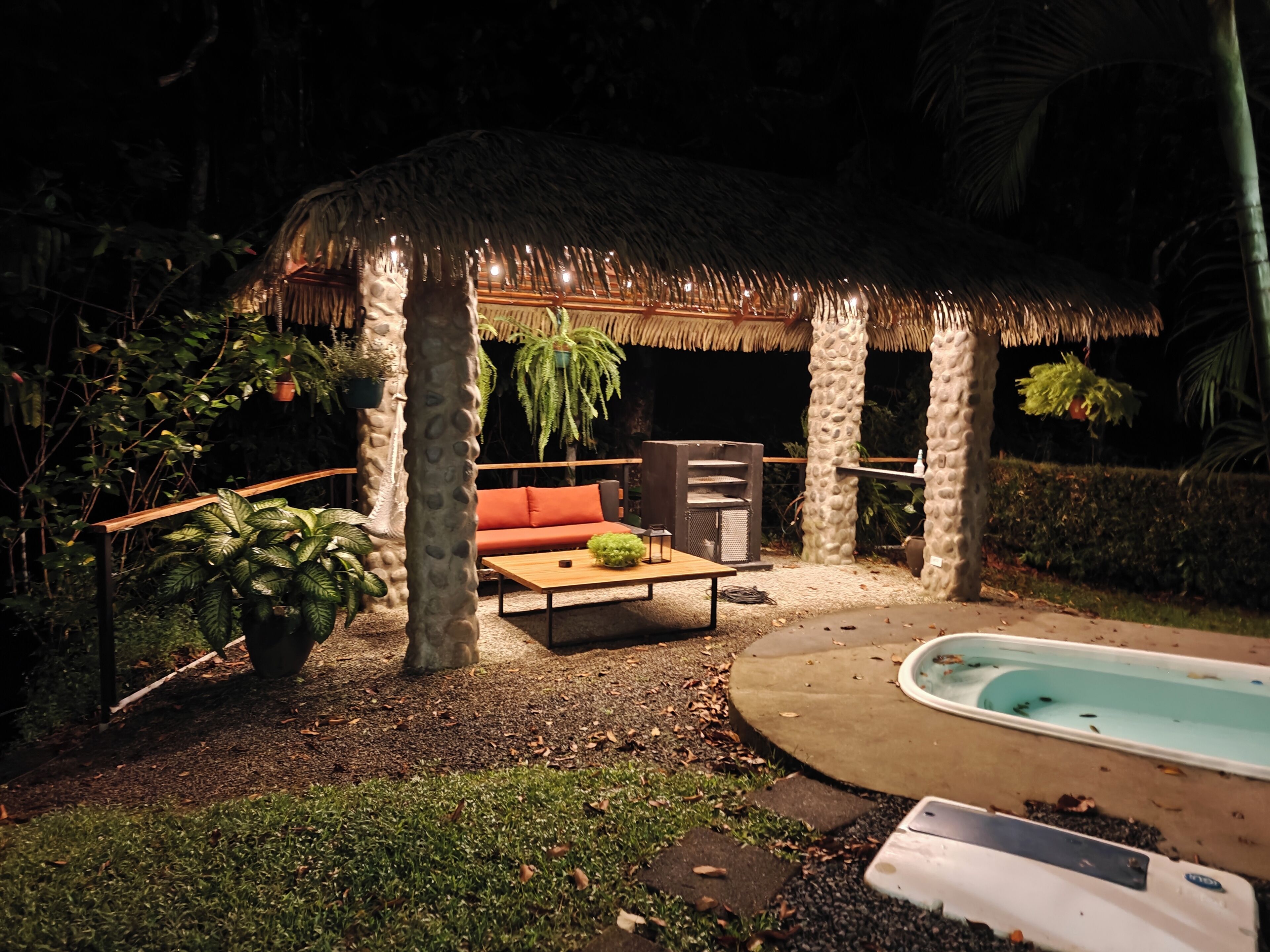 A thatched-roof outdoor seating area with a sofa and table, surrounded by tropical plants and a small pool.