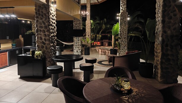 An outdoor patio area with stone pillars, round tables, and chairs, surrounded by potted plants and a pool.