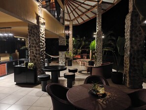 Ein Terrassenbereich im Freien mit Steinsäulen, runden Tischen und Stühlen, umgeben von Kübelpflanzen und einem Pool.