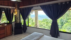 A room with a bed, a wooden dresser, a stone pillar, and a view of a mountainous landscape through large windows with dark curtains.