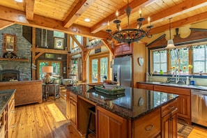 A spacious kitchen with a large island, wooden beams, stone fireplace, and a chandelier.