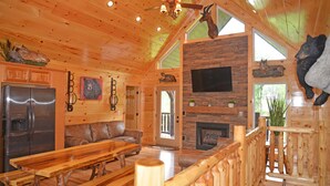 A wooden cabin interior with a stone fireplace, a large TV mounted above it, and a wooden bench and table in the center. - Luxury 4 King Suites, 4 Bath Log Cabin Close to Town & Trolley (Gatlinburg)