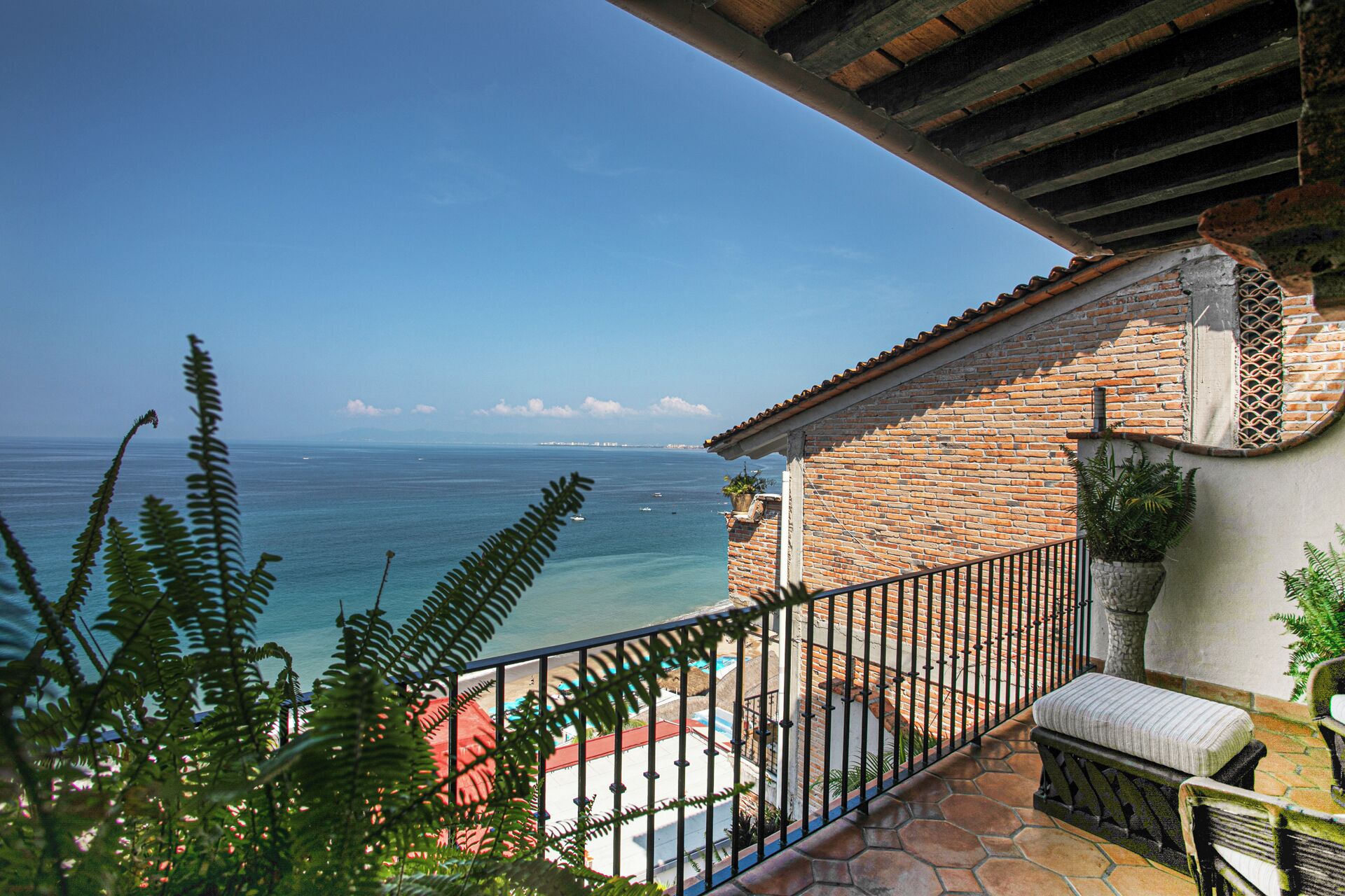 A balcony with a view of the sea, a brick building, and a stone patio with a bench.