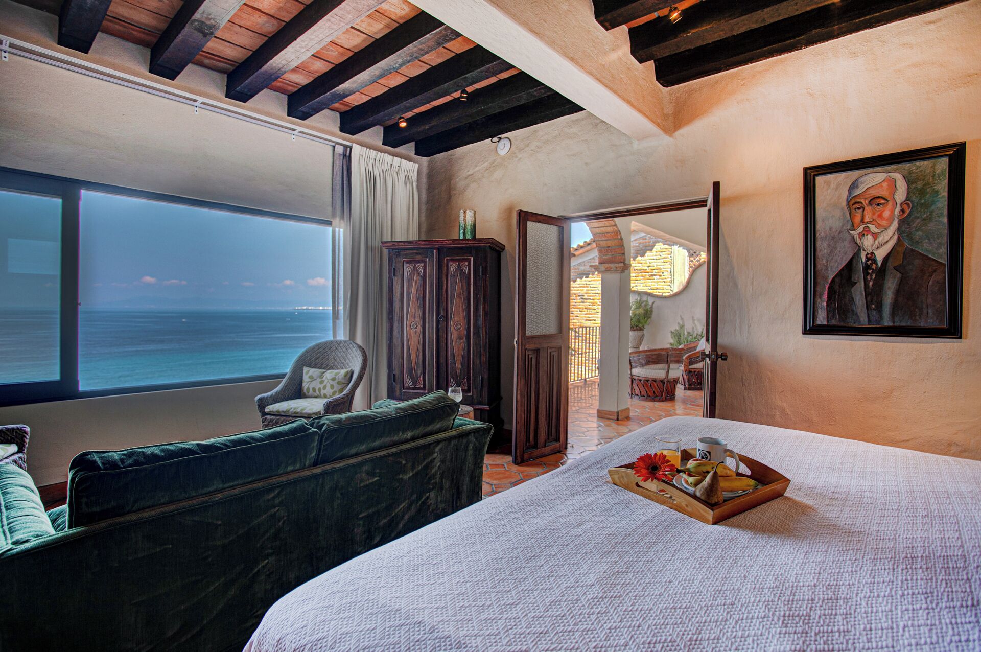 Une chambre avec une grande fenêtre donnant sur la mer, un lit avec un plateau de fruits et une tasse, une chaise et un meuble.