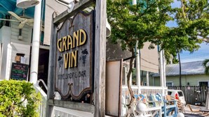 Ein hölzernes Schild für den Weinkeller und die Bar 'Grand Vin' mit weißer Veranda und Außenbestuhlung.