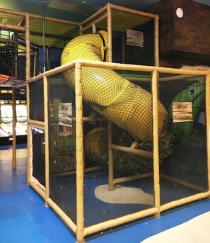 Un'ampia area recintata con uno scivolo a spirale giallo, staccionate di bambù e un cartello con la scritta 'NON NUTRIRE GLI ALLIGATORI'.