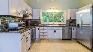 A kitchen with white cabinets, a stainless steel refrigerator, a toaster, and a window with a view of greenery. - Royal Palm: AC, Private Lanai, Walk to Poipu Beach (Koloa)