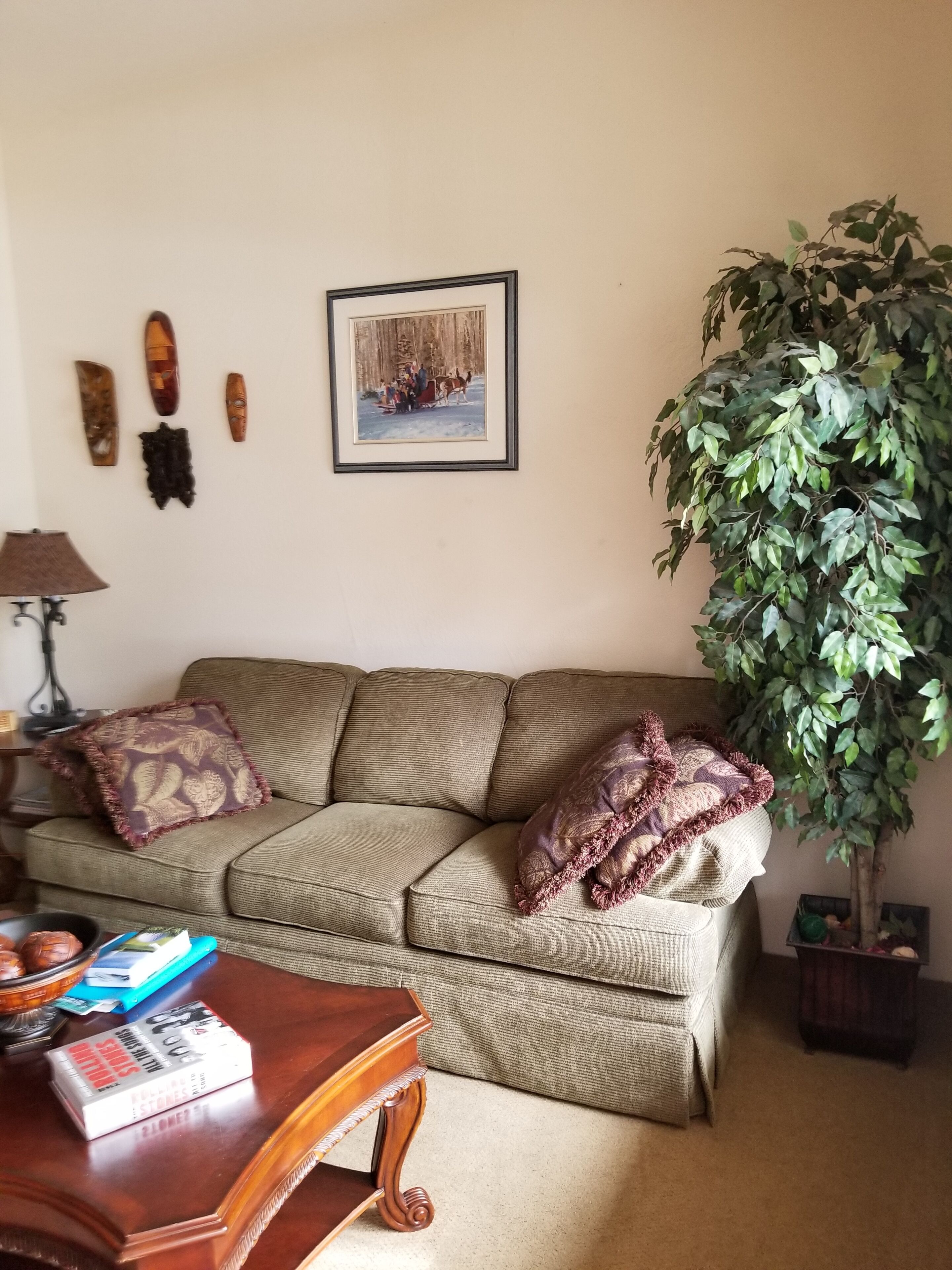 Ein Wohnzimmer mit einer Couch, einem Tisch, einer Lampe, einem gerahmten Bild und einer großen Pflanze.