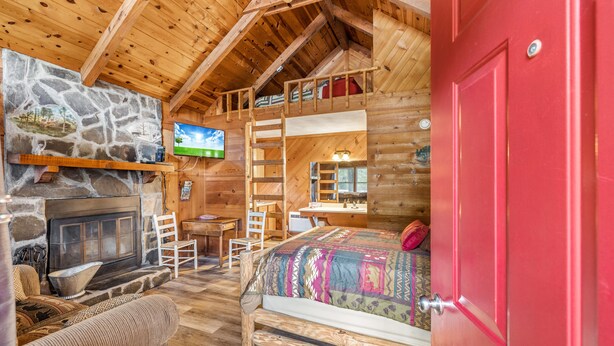 A cozy log cabin bedroom with a stone fireplace, a bunk bed, a desk, and a television.