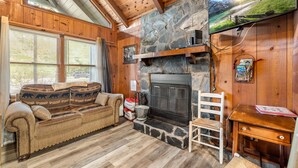 A cozy cabin interior with a stone fireplace, wooden walls, a sofa with patterned cushions, and a television mounted above it.